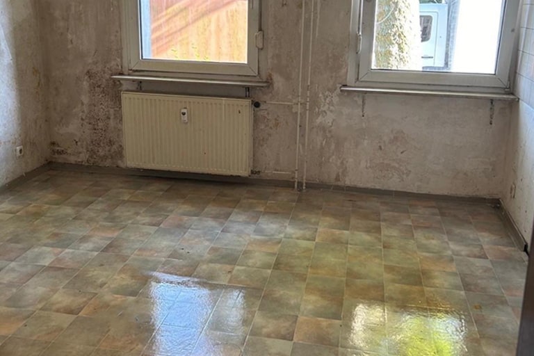 Ein leerer Raum mit abblätternder Farbe an den Wänden und zwei Fenstern. Der Boden besteht aus grün-beigen karierten Linoleumfliesen und unter einem Fenster ist ein an der Wand montierter Heizkörper zu sehen. Messie Küche entrümpeln und reinigen.