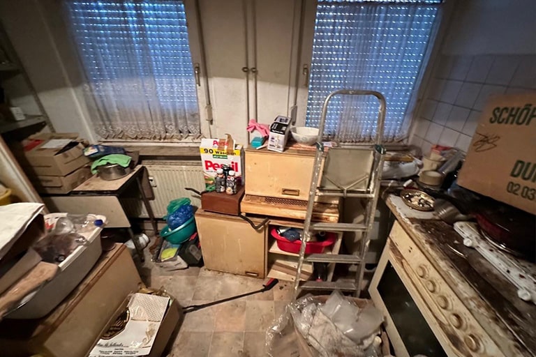 Eine überfüllte Küche mit überall verstreuten Kisten, Taschen und verschiedenen Gegenständen. Eine Leiter lehnt an einem Schrank. Im Hintergrund sind zwei Fenster mit transparenten Vorhängen zu sehen. Auf der rechten Seite ist ein alter Herd zu sehen. Reinigungsmittel und eine Packung Persil-Waschmittel liegen inmitten des Durcheinanders. Messie Küche entrümpeln und reinigen.